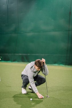 Erkek golfçü, tesisteki yeşil zemin ve golf eğitim ekipmanlarıyla çevrili, kapalı alandaki suni çimlere alıştırma yapmaya odaklanıyor.