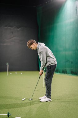 Genç adam kapalı bir golf sahasındaki suni sahaya golf topları koymaya odaklanıyor, yeşil ağlar ve geniş bir yerleşim alanı var.