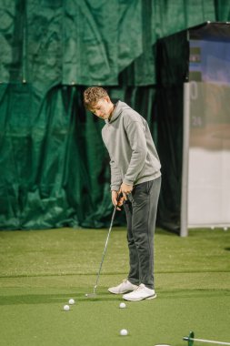 Genç erkek golfçü, suni çim ve görüş alanının geliştirilmesi için yeşil zemin içeren kapalı bir golf sahasında pratik yapmaya odaklanıyor.