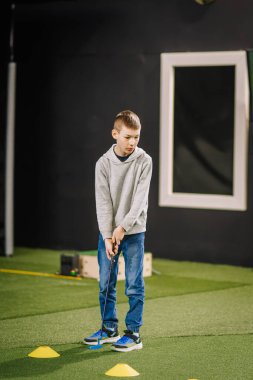Genç çocuk, arka planda yapay çim ve çeşitli eğitim araçlarının yer aldığı bir golf sahasının içinde pratik yapmaya odaklanıyor.