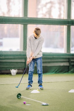 Genç çocuk, arka planda yapay çim ve çeşitli eğitim araçları bulunan kapalı bir golf sahasında golf salıncağına odaklanıyor.