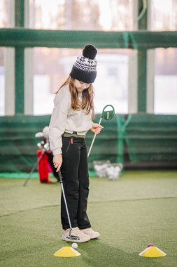 Kışlık şapkalı genç kız içeride golf oynuyor, sentetik çimlerin üzerinde duruyor, hizalama ve rehberlik için renkli işaretlerle.