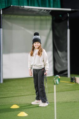 Genç kız kapalı bir golf sahasında yeşil bir alanda duruyor. Koniler ve antrenman için yakınlarda bir golf sopası var.