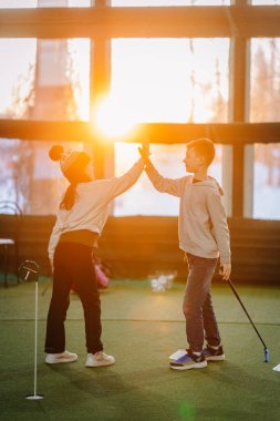 İki çocuk kapalı alanda golf oynarken, doğal ışık sağlayan suni çim ve geniş pencerelerle çevrili bir beşlik çakıyor.