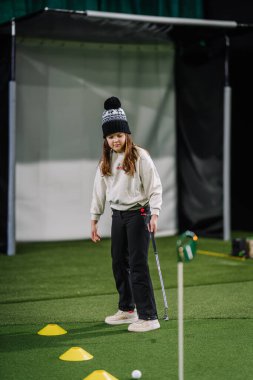 Genç kız içeride golf antrenmanına odaklanır, günlük kıyafetler giyer, arka planda koni gibi bir yere ve eğitim yardımcılarına sahiptir.