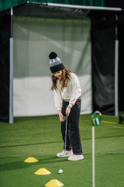 Genç kız antrenman sahasında golf topunu içeri sokmaya odaklanıyor, kışlık şapka takıyor, yeşil çimenlikte eğitim konileri düzenleniyor.