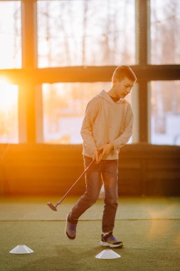 Genç çocuk antrenman sahasında golf salıncağına odaklanıyor. Suni çim ve koniler sunuyor. Güneş ışığı uzayı aydınlatıyor.
