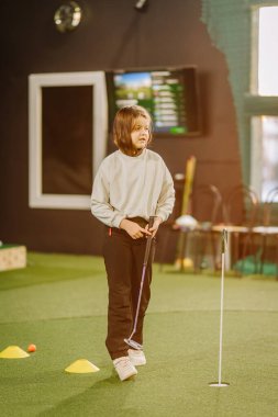 Genç bir çocuk, elinde eğitim araçları ve arka planda dijital ekranı olan bir golf sopası tutarak yeşil bir alanda duruyor.