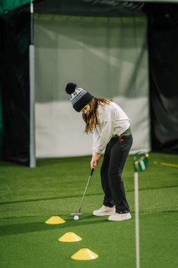 Genç bir kız golf topunu yeşil bir alana yerleştiriyor. Önünde de antrenman konileri var. Golf tekniğini geliştiriyor.