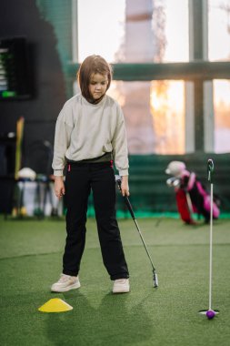 Genç bir kız suni çimlerin üzerinde durur ve arka planda golf ekipmanları olan bir eğitim konisine konsantre olur.