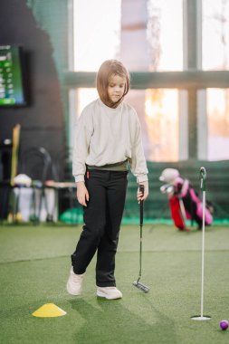 Genç bir kız suni çayırda duruyor. Tekniğini içeride uyguluyor. Eğitim araçları ve arka planda görünen bir golf deliği var.