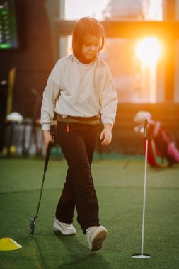 Genç kız içeride golf oynarken hedefine doğru yürüyor, güneş ışığı geniş pencerelerden boşluğu aydınlatıyor.