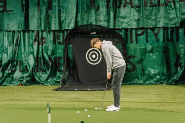 Erkek golfçü, hedef ağ ve yapay yeşil çim yüzeyi içeren kapalı bir golf eğitim alanında pratik yapmaya odaklanır.