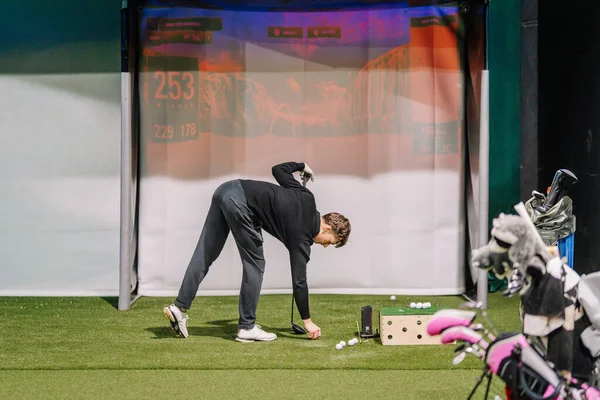 Erkek golfçü, yakın çevresindeki ölçümleri ve golf sopalarını gösteren dijital bir ekranla kapalı alanda golf topunu atmak için eğilir.