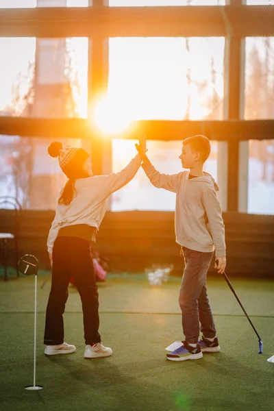 İki çocuk kapalı alanda golf oynarken, doğal ışık sağlayan suni çim ve geniş pencerelerle çevrili bir beşlik çakıyor.