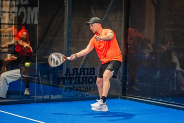 Riga, Letonya - 24 Ocak 2026: Padel turnuvası. Turuncu tişörtlü ve siyah şortlu bir erkek sporcu mavi bir padel kortunda raketle topa vuruyor.