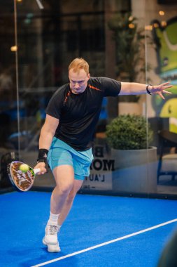 Riga, Letonya - 24 Ocak 2026: Padel turnuvası. Siyah tişörtlü ve mavi şortlu erkek padel oyuncu mavi kortta camlarla çevrili bir raket sallıyor.