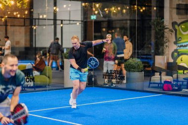 Riga, Letonya - 24 Ocak 2026: Padel turnuvası. Siyah tişörtlü ve mavi şortlu erkek padel oyuncusu arka planda izleyicilerle birlikte salıncakta sallanıyor.