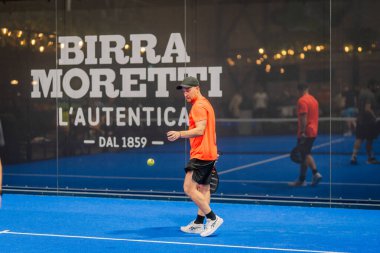 Riga, Letonya - 24 Ocak 2026: Padel turnuvası. Parlak turuncu tişörtlü erkek padel oyuncusu mavi kortta duruyor. Arka planda Birra Moretti logosu olan topa vurmaya hazır.