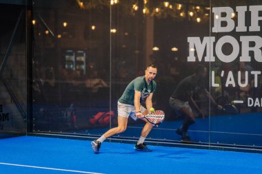 Riga, Letonya - 24 Ocak 2026: Padel turnuvası. Yeşil tişörtlü ve şortlu erkek padel oyuncusu mavi kortta camdan duvarları olan topa vurmaya hazırlanıyor.