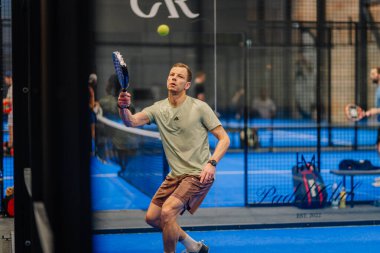 Riga, Letonya - 24 Ocak 2026: Padel turnuvası. Açık tişörtlü ve şortlu bir erkek sporcu, mavi yüzeyi olan kapalı bir padel kortunda topa vurmaya hazırlanıyor.