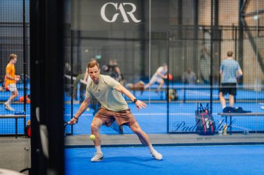 Riga, Letonya - 24 Ocak 2026: Padel turnuvası. Açık tişörtlü ve şortlu erkek padel oyuncusu diğer oyuncularla birlikte mavi sahada topa vurmaya hazırlanıyor.