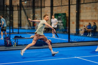 Riga, Letonya - 24 Ocak 2026: Padel turnuvası. Açık tişörtlü ve şortlu bir erkek sporcu, mavi bir padel kortunda raketle topa vuruyor.