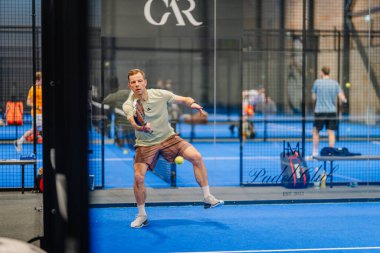 Riga, Letonya - 24 Ocak 2026: Padel turnuvası. Gri tişörtlü ve şortlu erkek sporcu, arka planda oyuncularla birlikte mavi bir padel kortunda topa vurmaya hazırlanıyor.