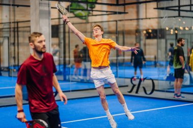 Riga, Letonya - 24 Ocak 2026: Padel turnuvası. Turuncu tişörtlü genç erkek padel oyuncu, mavi sahada başka bir oyuncu hazır dururken topa vurmak için zıplıyor.