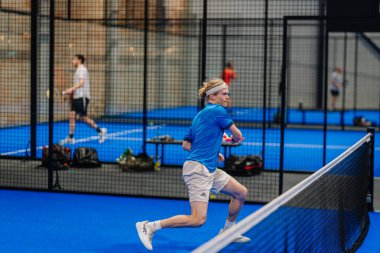 Riga, Letonya - 24 Ocak 2026: Padel turnuvası. Mavi tişörtlü ve şortlu genç erkek padel oyuncu, kapalı alanda mavi bir sahada şort sallıyor.