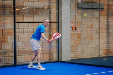Riga, Letonya - 24 Ocak 2026: Padel turnuvası. Mavi tişörtlü ve şortlu erkek padel oyuncusu mavi kortta kırmızı raketle topa vurmaya hazırlanıyor.
