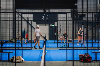 Riga, Letonya - 24 Ocak 2026: Padel turnuvası. Beyaz tişörtlü ve gri şortlu erkek padel oyuncusu mavi bir kortta seyirciler ve ağlarla yürüyor.