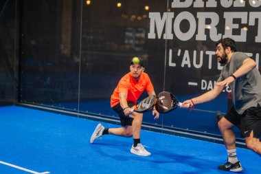 Riga, Letonya - 24 Ocak 2026: Padel turnuvası. İki adam mavi kortta Padel oynuyor, biri turuncu tişörtlü, diğeri gri gömlekli, arkasında da cam bir duvar var.