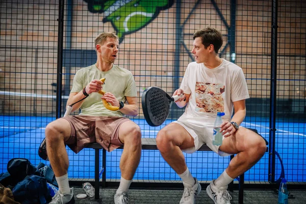 Riga, Letonya - 24 Ocak 2026: Padel turnuvası. İki erkek Padel oyuncusu bir bankta rahatlıyor, biri bir muz tutuyor ve diğeri de bir su şişesi, spor tesisinde.
