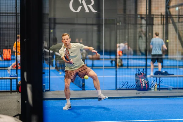 Riga, Letonya - 24 Ocak 2026: Padel turnuvası. Gri tişörtlü ve şortlu erkek sporcu, arka planda oyuncularla birlikte mavi bir padel kortunda topa vurmaya hazırlanıyor.