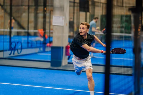 Riga, Letonya - 24 Ocak 2026: Padel turnuvası. Siyah tişörtlü ve beyaz şortlu erkek padel oyuncusu, mavi kortta cam duvarlı topa vuruyor.
