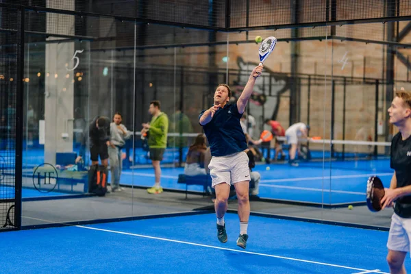 Riga, Letonya - 24 Ocak 2026: Padel turnuvası. Mavi tişörtlü erkek padel oyuncu, mavi kortta raketle topa vurmak için atlıyor.