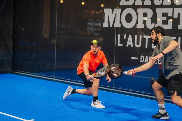Riga, Letonya - 24 Ocak 2026: Padel turnuvası. İki adam mavi kortta Padel oynuyor, biri turuncu tişörtlü, diğeri gri gömlekli, arkasında da cam bir duvar var.