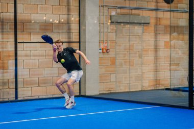Riga, Letonya - 24 Ocak 2026: Padel turnuvası. Siyah tişörtlü ve beyaz şortlu erkek padel oyuncusu mavi sahada camdan duvarlarla topa vurmaya hazırlanıyor.