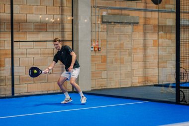 Riga, Letonya - 24 Ocak 2026: Padel turnuvası. Siyah tişörtlü ve beyaz şortlu erkek padel oyuncusu mavi kortta camdan duvarlı bir topa vurmaya hazırlanıyor.