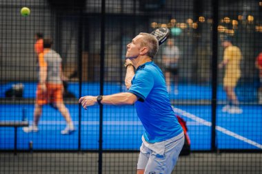 Riga, Letonya - 24 Ocak 2026: Padel turnuvası. Mavi tişörtlü erkek padel oyuncu, kapalı alanda mavi yüzeyi olan bir sahada oynanan maçta topa vurmaya odaklanıyor.