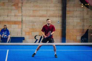 Riga, Letonya - 24 Ocak 2026: Padel turnuvası. Kırmızı tişörtlü erkek padel oyuncu mavi sahada topa vurmaya hazır.