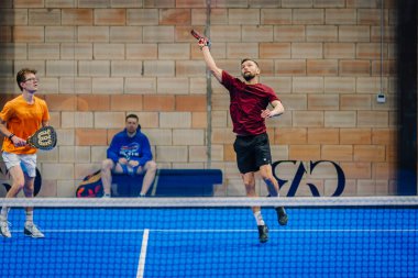 Riga, Letonya - 24 Ocak 2026: Padel turnuvası. Kırmızı tişörtlü erkek atlet, turuncu tişörtlü takım arkadaşı iç sahada oynamaya hazırlanırken padel topu servis ediyor.