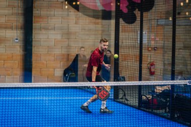 Riga, Letonya - 24 Ocak 2026: Padel turnuvası. Kırmızı tişörtlü erkek padel oyuncu, arka planda seyircilerin olduğu mavi sahada topa vurmaya hazır.