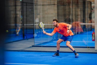Riga, Letonya - 24 Ocak 2026: Padel turnuvası. Turuncu tişörtlü ve mavi şortlu erkek padel oyuncusu mavi kortta cam duvarlı bir topa vurmaya hazırlanıyor.