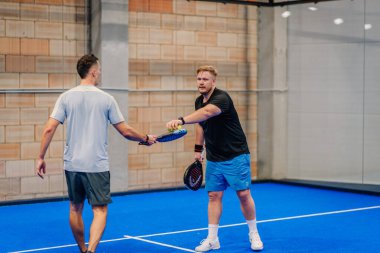 Riga, Letonya - 24 Ocak 2026: Padel turnuvası. Spor kıyafetleri içinde iki adam mavi bir kortta geniş bir tasarımı olan kapalı bir tesiste padel raketlerini teslim etti.