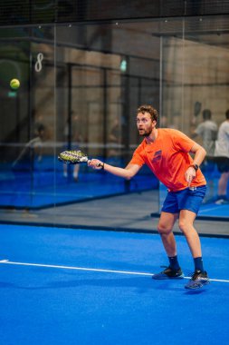 Riga, Letonya - 24 Ocak 2026: Padel turnuvası. Turuncu tişörtlü ve mavi şortlu erkek padel oyuncusu mavi kortta camdan duvarlarla topa vurmaya hazırlanıyor.