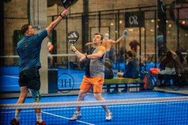 Riga, Letonya - 24 Ocak 2026: Padel turnuvası. İki adam mavi kortta Padel oynuyor, biri topa vurmaya hazırlanırken diğeri arka planda seyirciler var.