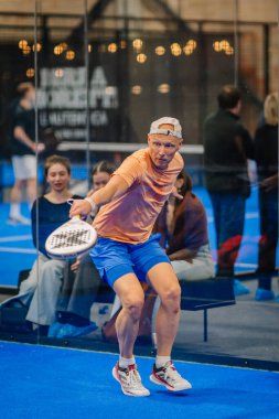 Riga, Letonya - 24 Ocak 2026: Padel turnuvası. Turuncu ve mavi giysili erkek oyuncu, kapalı sahada topa vuracak pozisyonda ve seyirciler izliyor.