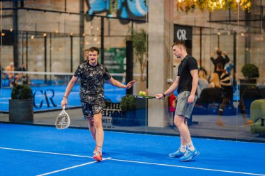 Riga, Letonya - 24 Ocak 2026: Padel turnuvası. İki erkek Padel oyuncusu mavi kortta antrenman yapıyor, biri raket tutarken diğeri spor tesisinde çalışmaya hazırlanıyor.